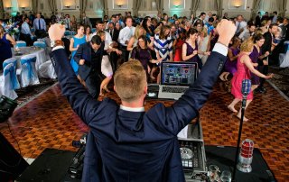 A DJ raises his arms in front of a lively crowd dancing at an indoor event with a wooden floor.