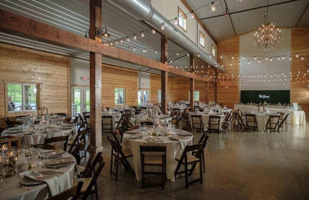 Spacious wooden reception hall with round tables set for an event. String lights hang from the ceiling, and a chandelier is visible.