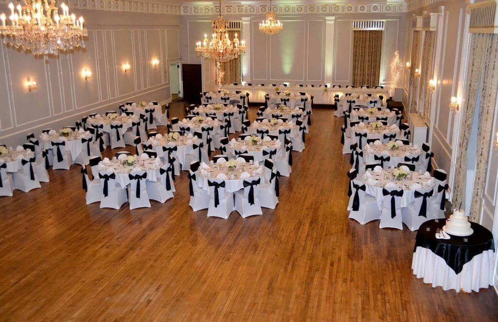 Ballroom with wooden floor, set for an event with round tables covered in white cloths and black bows. Decor includes chandeliers and a tiered cake on a separate table.