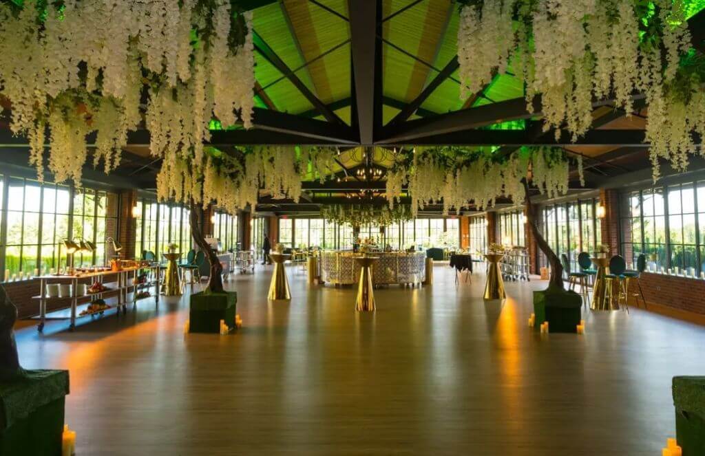 Spacious, elegant event hall with hanging white flowers, large windows, and a central table. Several smaller tables and seats are arranged throughout the wooden-floored room.