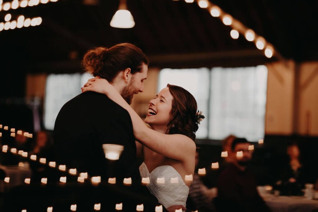 A bride and groom dance together indoors, smiling at each other. Twinkling string lights and out-of-focus guests are visible in the background, creating a warm atmosphere.