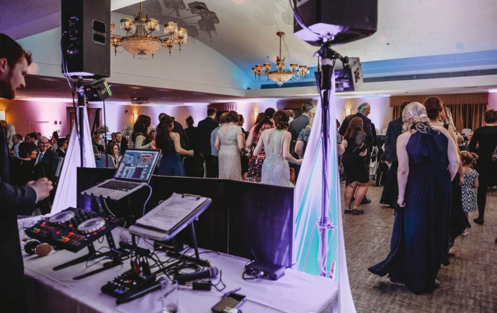 A DJ booth with equipment is set up in the foreground at an indoor event. Groups of people are standing and mingling on a carpeted dance floor under chandeliers and soft, colorful lighting.