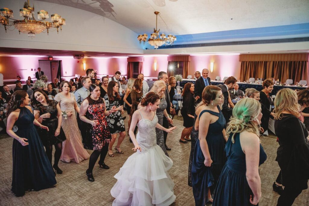 A group of people, including a bride in a white gown, dance together in a decorated banquet hall with chandeliers, pink-lit walls, and guests seated at tables in the background.