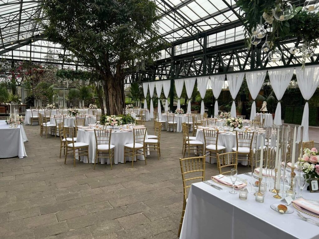 A spacious indoor venue with a glass roof is set up for an event, featuring round tables with white tablecloths, gold chairs, floral centerpieces, and draped white curtains along the walls.