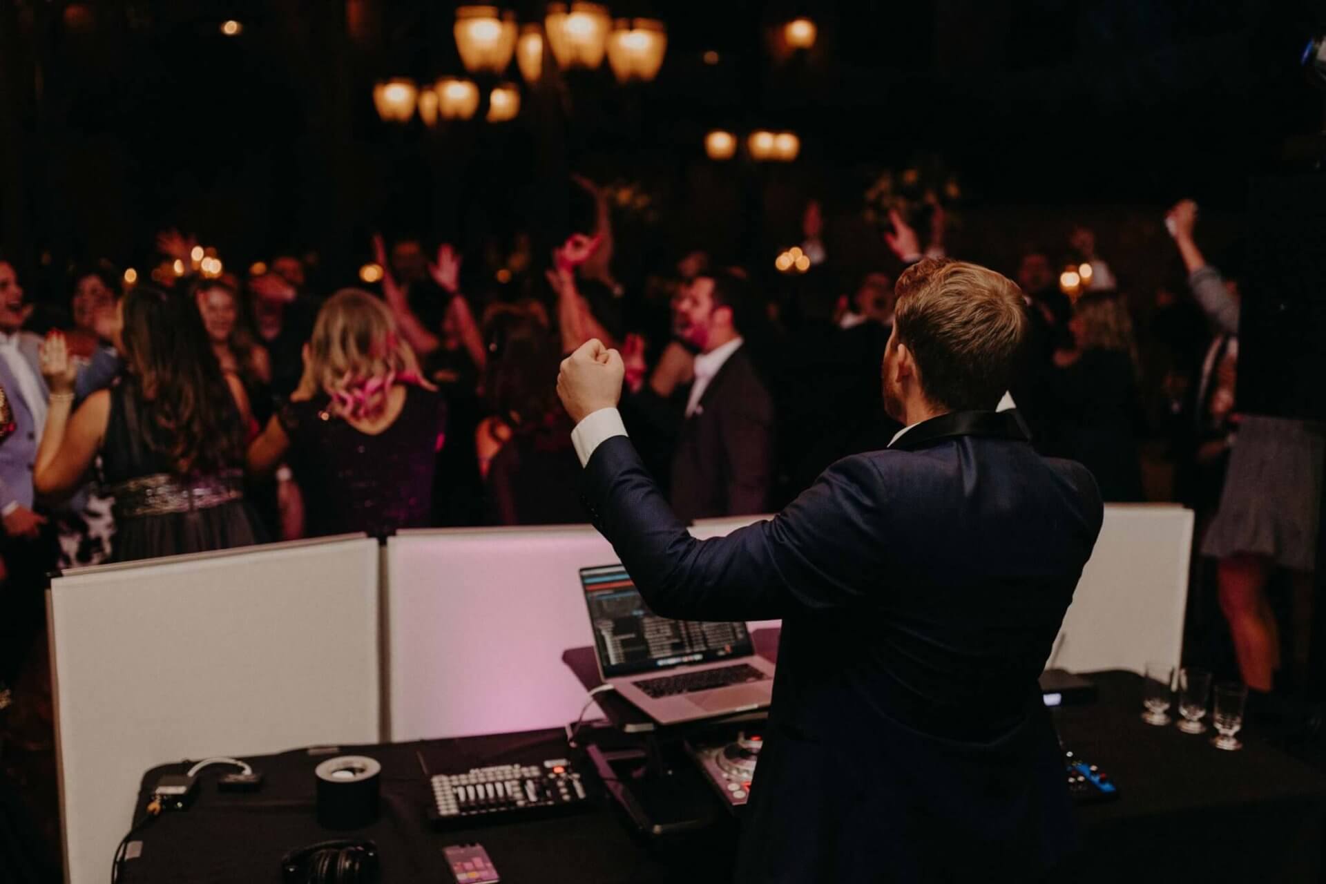 A DJ in a suit is playing music at a lively party, with guests dancing and raising their hands. The room is dimly lit with chandeliers overhead.