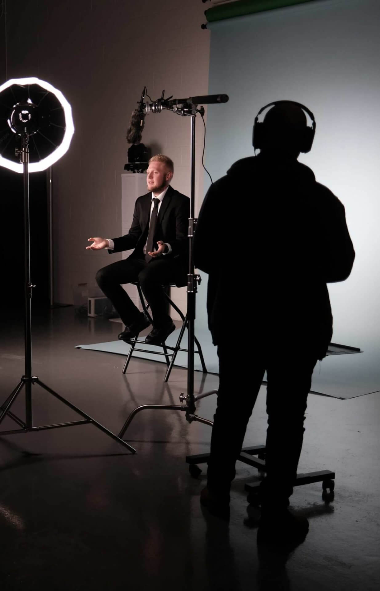 commercial-videography-michaels-entertainment A person in a suit sits under studio lighting while another stands nearby with headphones, holding a microphone boom.