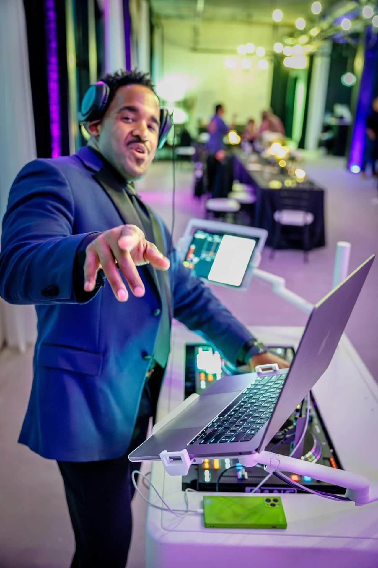 A DJ wearing headphones is pointing towards the camera while standing at a mixing desk with a laptop and other equipment in a colorful room.