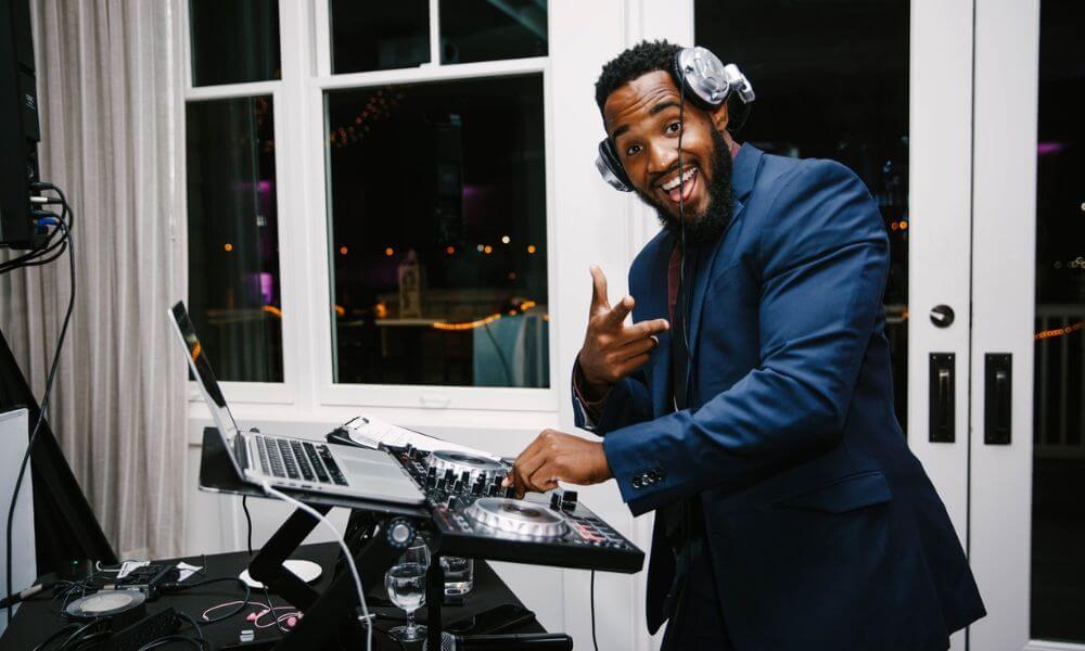 DJ wearing headphones, smiling and pointing while mixing music on a DJ controller with a laptop on a stand.