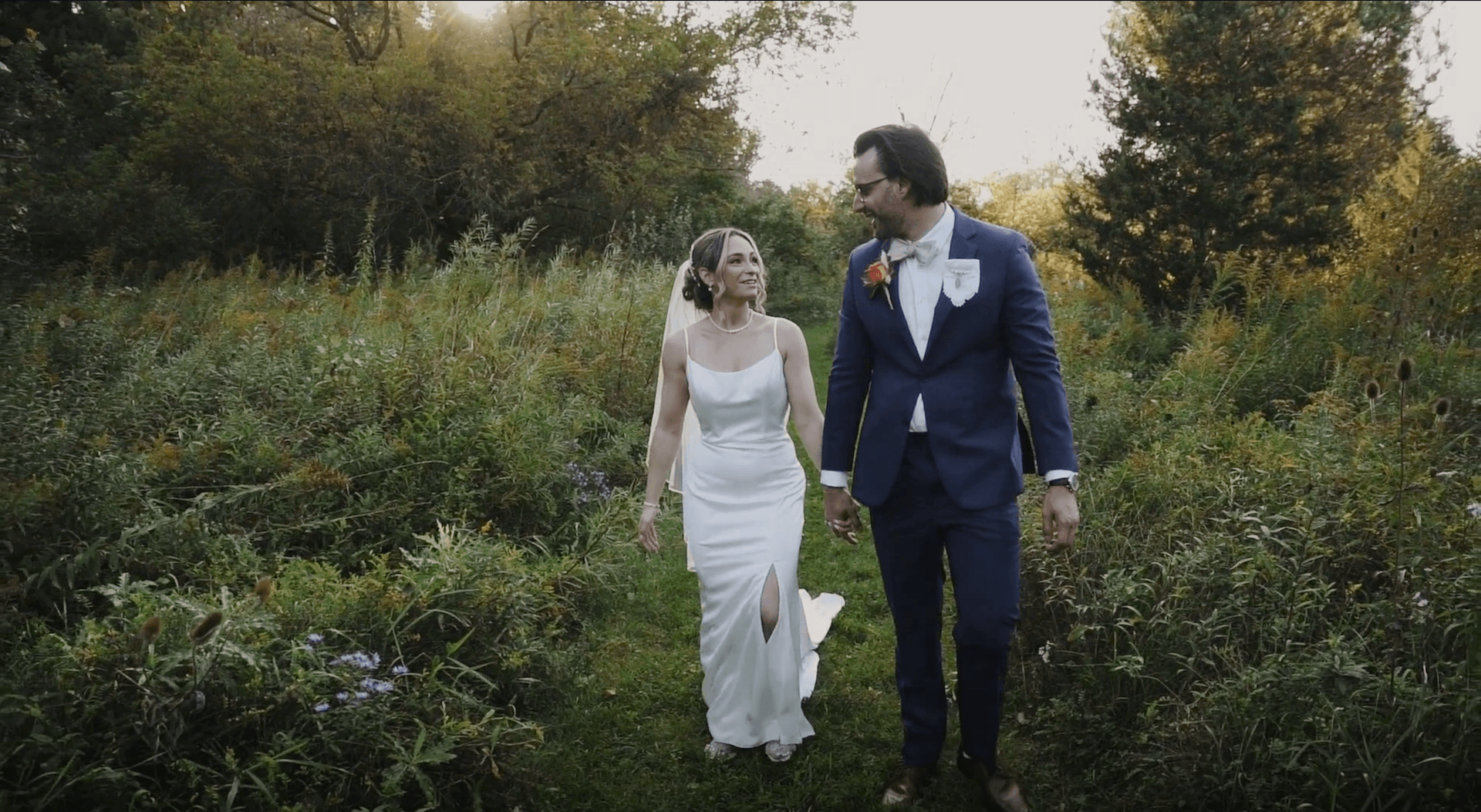A bride and groom walk hand in hand through a grassy, wooded area. She wears a white dress with a slit, and he wears a blue suit with a red boutonniere. Trees and sunlight are in the background.