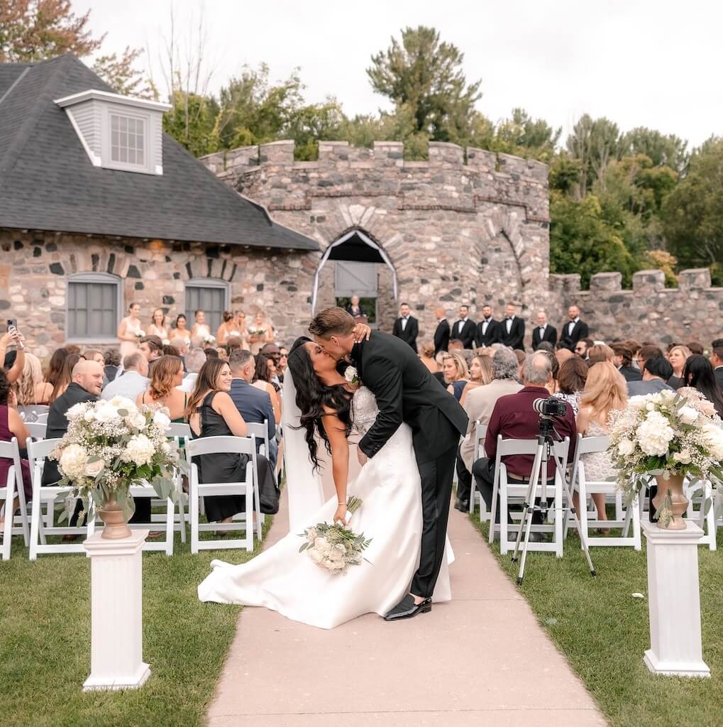 A couple kisses at an outdoor wedding ceremony in front of guests, near a stone building with arches, surrounded by greenery.