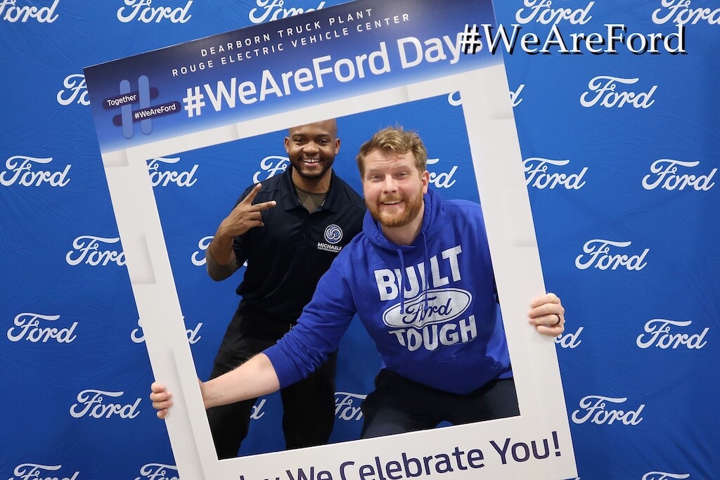 Two men pose and smile inside a large hashtag-themed photo frame in front of a blue Ford-branded backdrop at a Ford event.