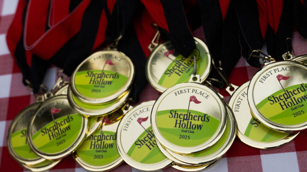A pile of gold medals with black ribbons, labeled “First Place Shepherds Hollow Golf Club 2025,” displayed on a red and white checkered surface, shines as Michaels-Entertainment-Brings-Smiles-to-Special Olympics celebrations.