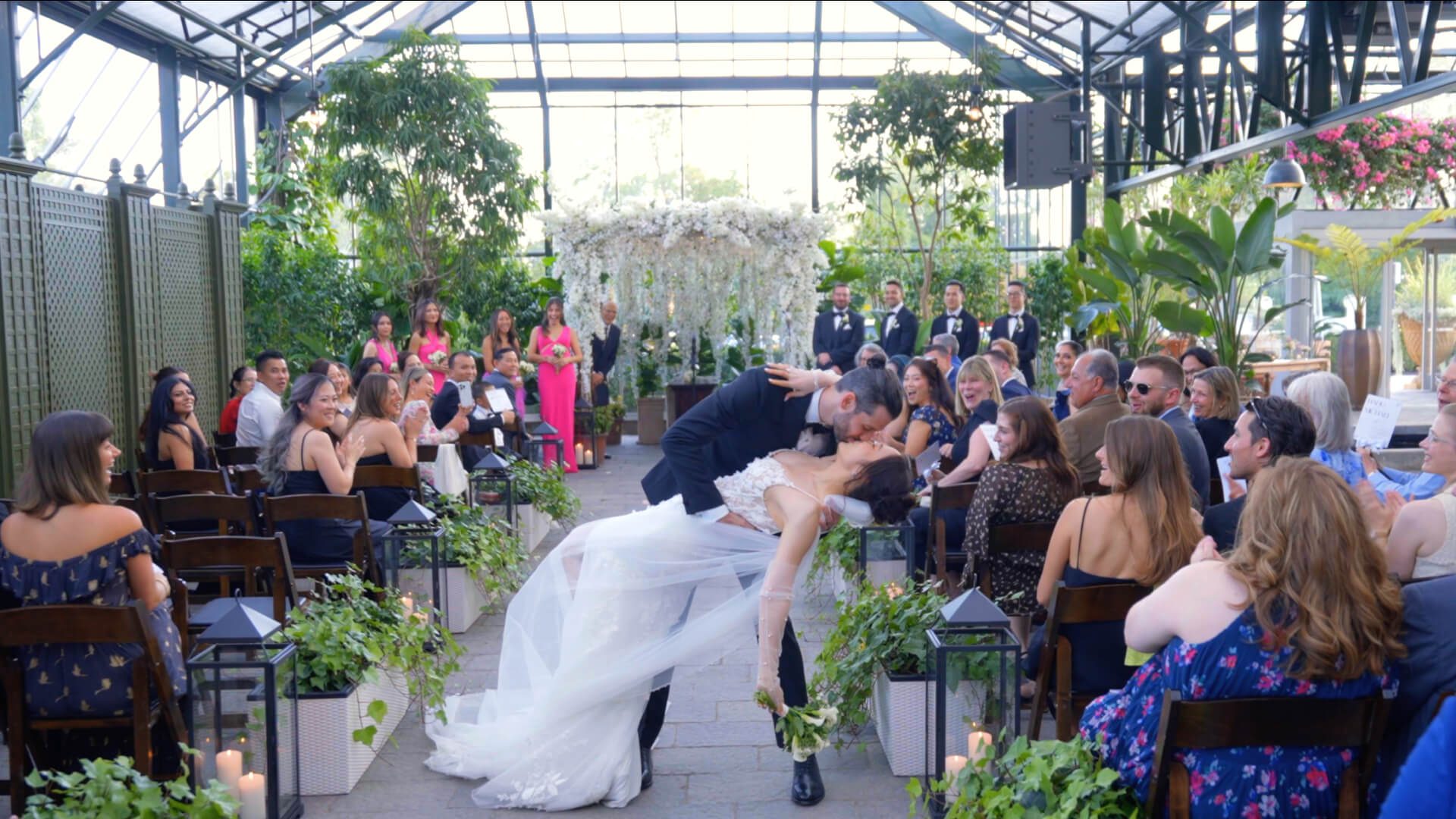 A Detroit bride and groom share a dramatic kiss at the end of the aisle during a wedding ceremony in a greenhouse, surrounded by seated guests and abundant greenery.