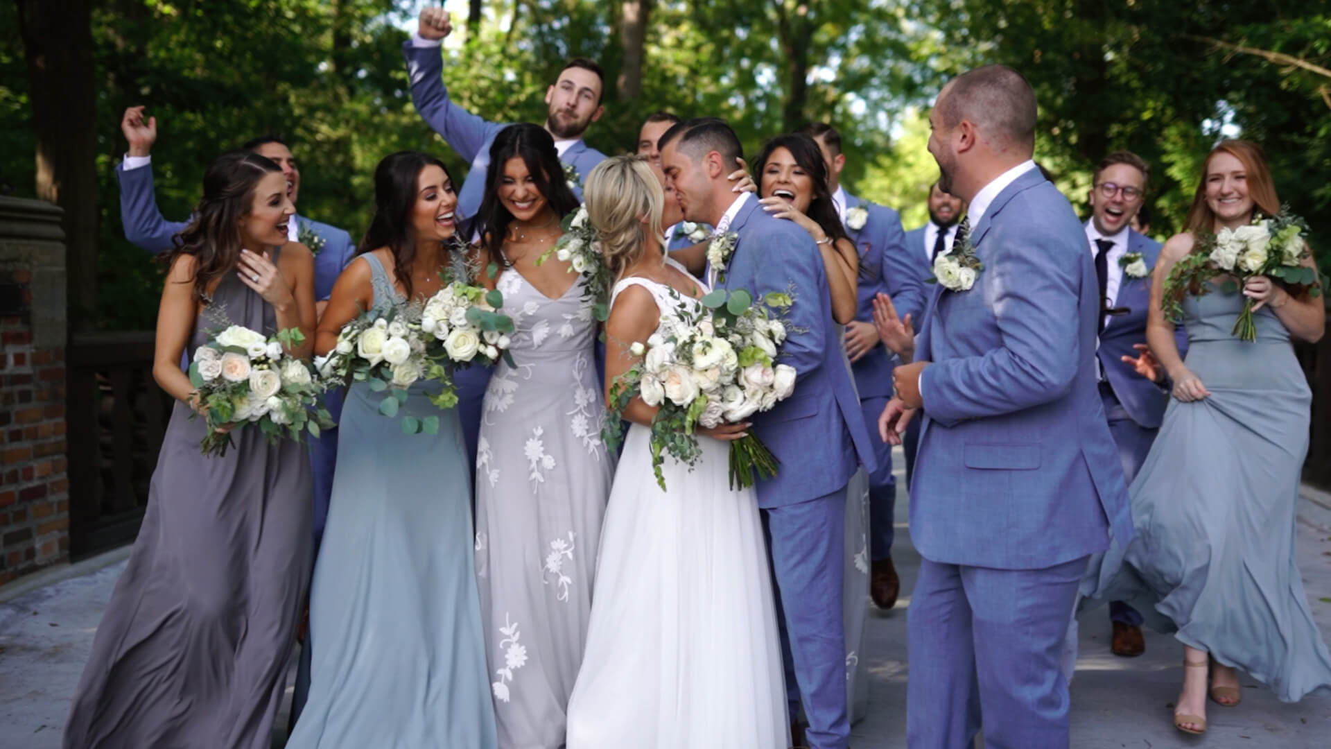 A Michigan wedding party, including bridesmaids in lavender dresses and groomsmen in blue suits, celebrates outdoors on a sunny day. The bride and groom stand at the center, holding bouquets and smiling, surrounded by friends.