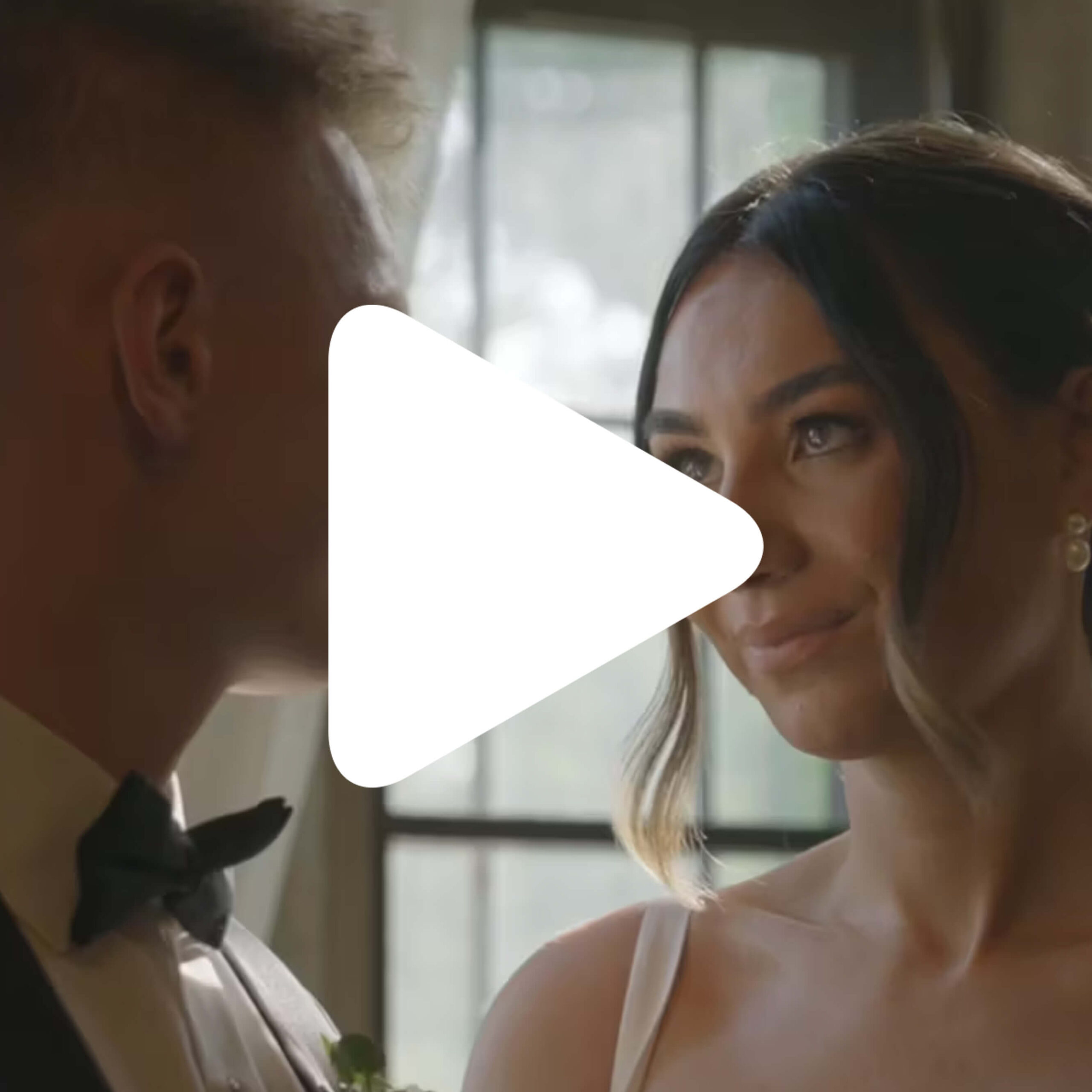 A bride and groom stand close to each other indoors, looking at each other, with natural light coming through a window behind them. A large white play button icon is overlaid in the center.