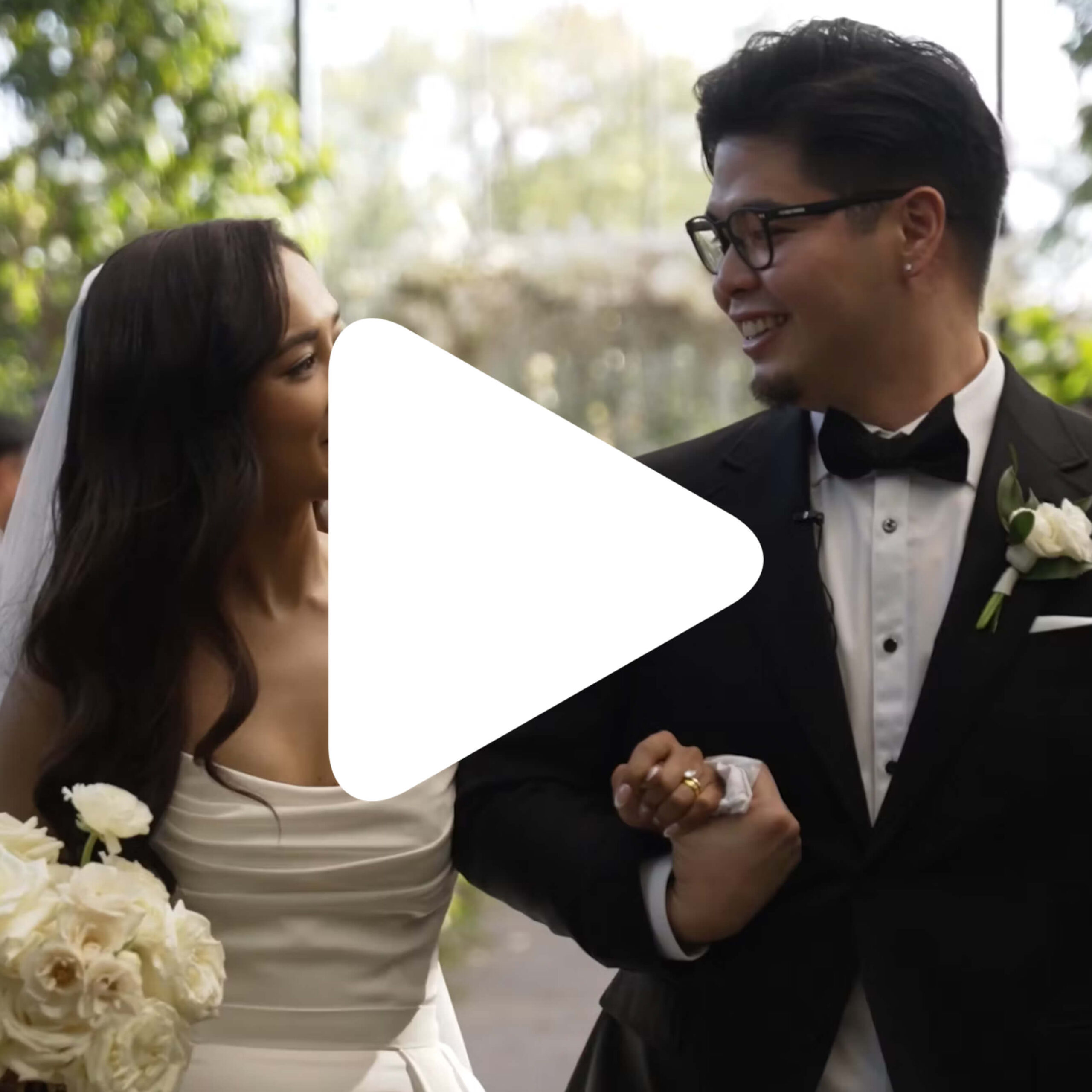A bride in a white dress holding a bouquet and a groom in a black tuxedo walk arm in arm, smiling at each other. There is a large play button icon over the center of the image.