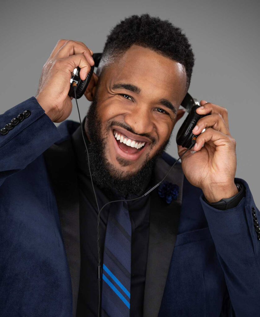 A man wearing a navy blue suit and striped tie smiles while holding a pair of headphones to his ears, against a plain grey background.