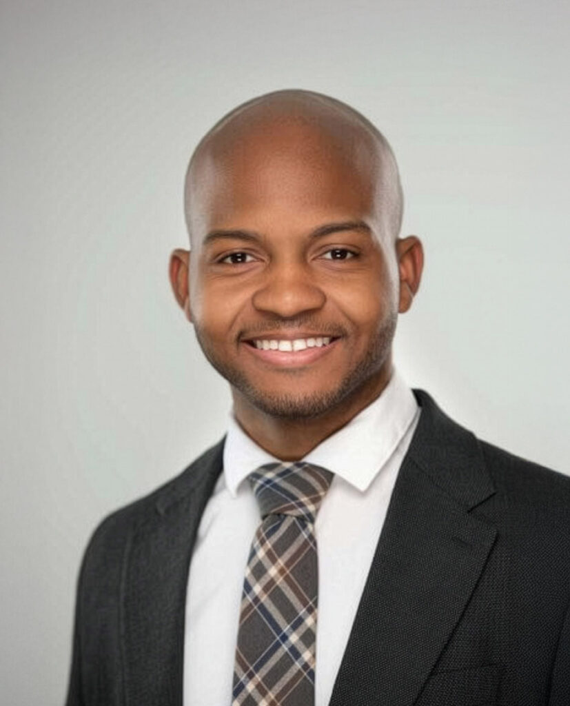 A man wearing a dark suit, white shirt, and plaid tie is smiling at the camera against a plain light gray background.