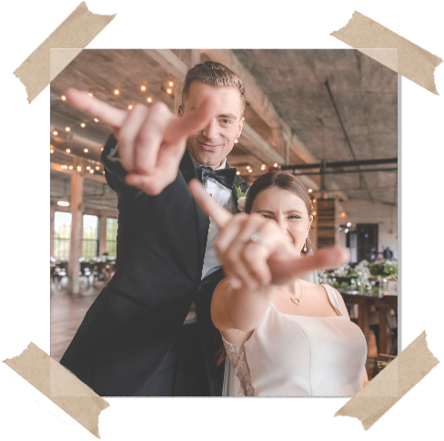 A bride and groom indoors, both smiling and making hand gestures toward the camera, with string lights and tables visible in the background. The image has a taped border effect.