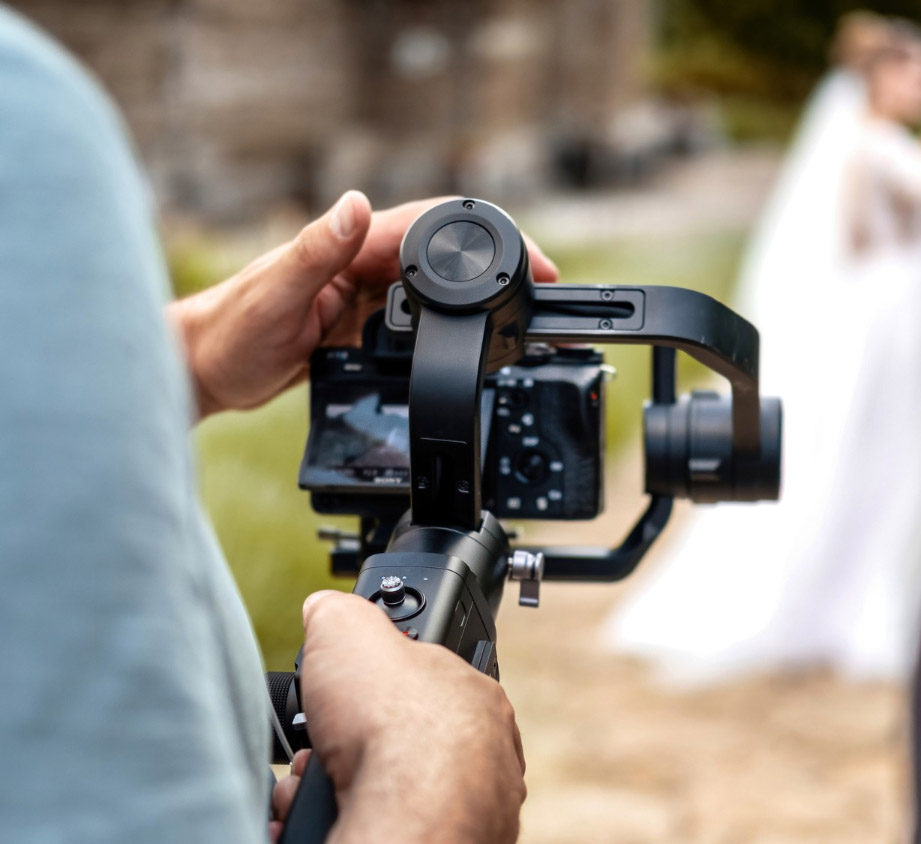 A person operates a handheld camera stabilizer outdoors, filming a scene with a woman in a white dress and veil in the blurred background.