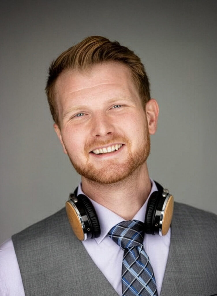 A man with reddish hair and a beard is smiling at the camera. He is wearing a gray vest, patterned tie, light shirt, and has headphones resting around his neck. The background is plain and gray.