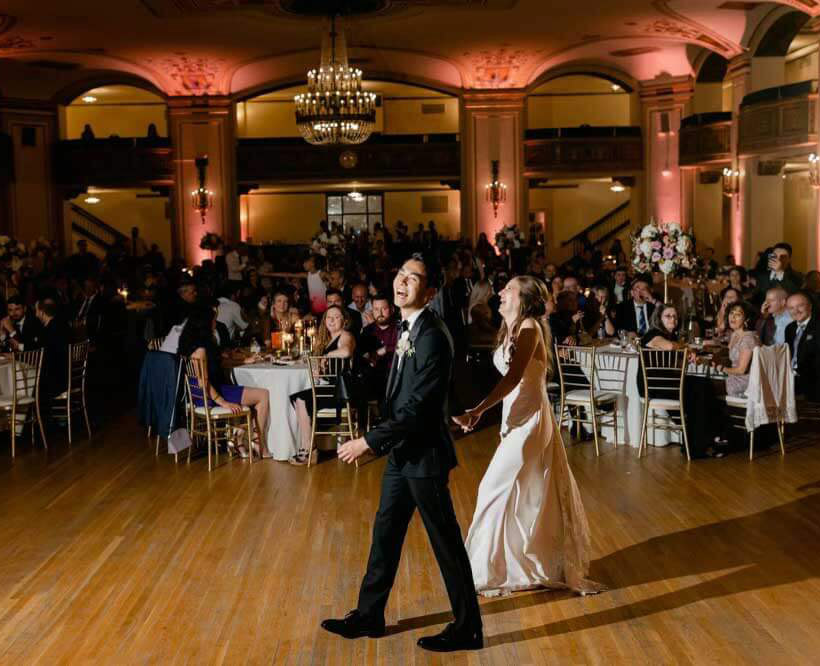 A bride and groom walk hand in hand across a ballroom dance floor, smiling, with guests seated at round tables in the background under warm lighting.