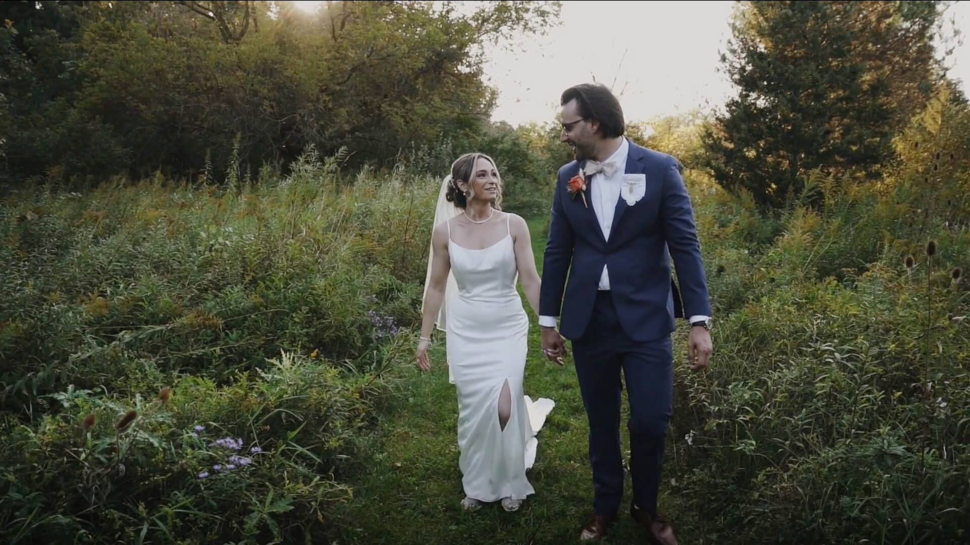 A bride in a white dress and a groom in a blue suit walk hand in hand through a grassy field surrounded by greenery and trees, looking at each other and smiling.