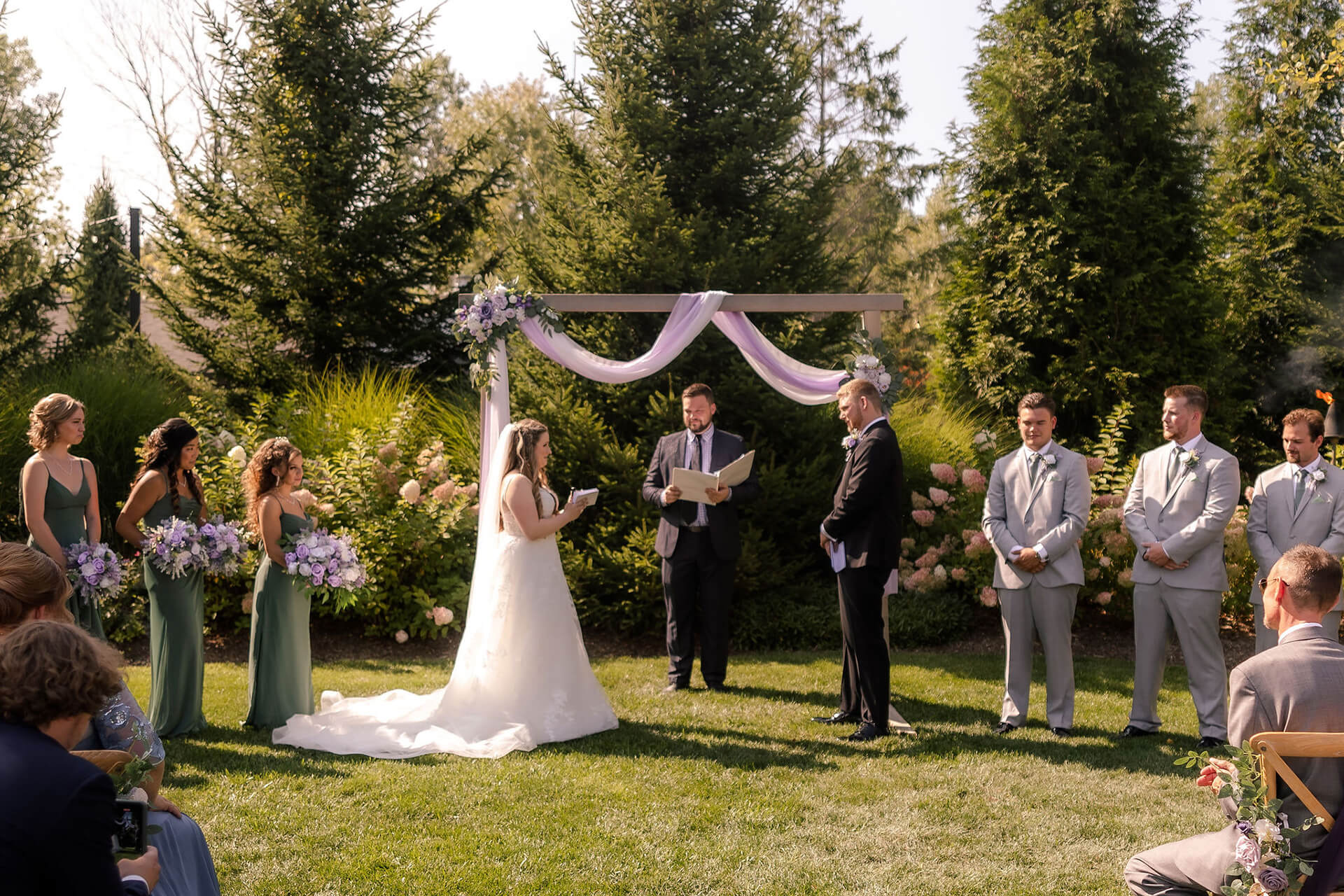 A bride and groom stand under a decorated wooden arch during an outdoor wedding ceremony. Bridesmaids in green dresses and groomsmen in gray suits stand nearby. The officiant reads from a book. Guests are seated on the lawn.