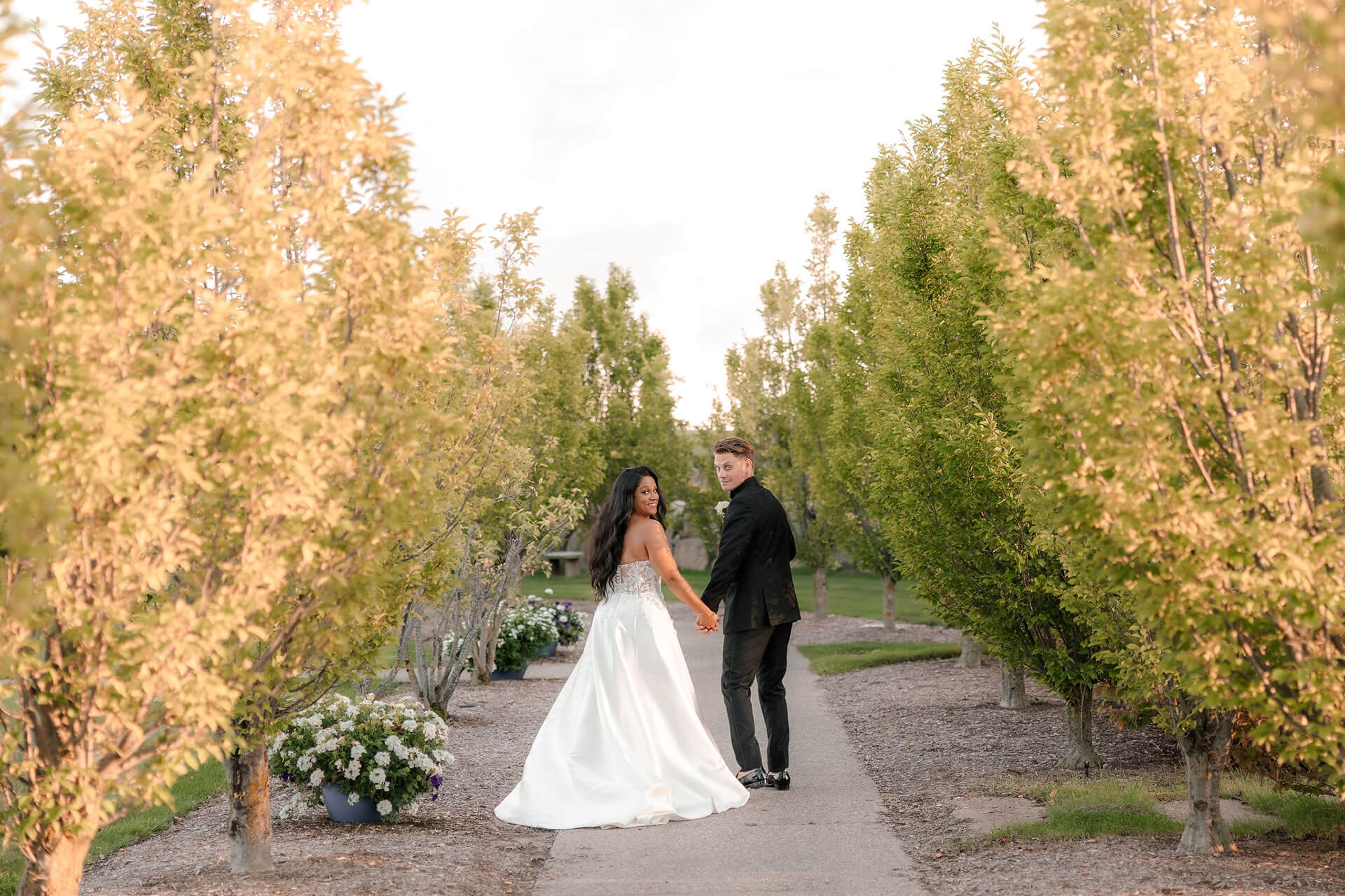 A bride in a white dress and a groom in a black suit walk hand in hand down a tree-lined path, both looking back toward the camera. The scene is bright and surrounded by green foliage.