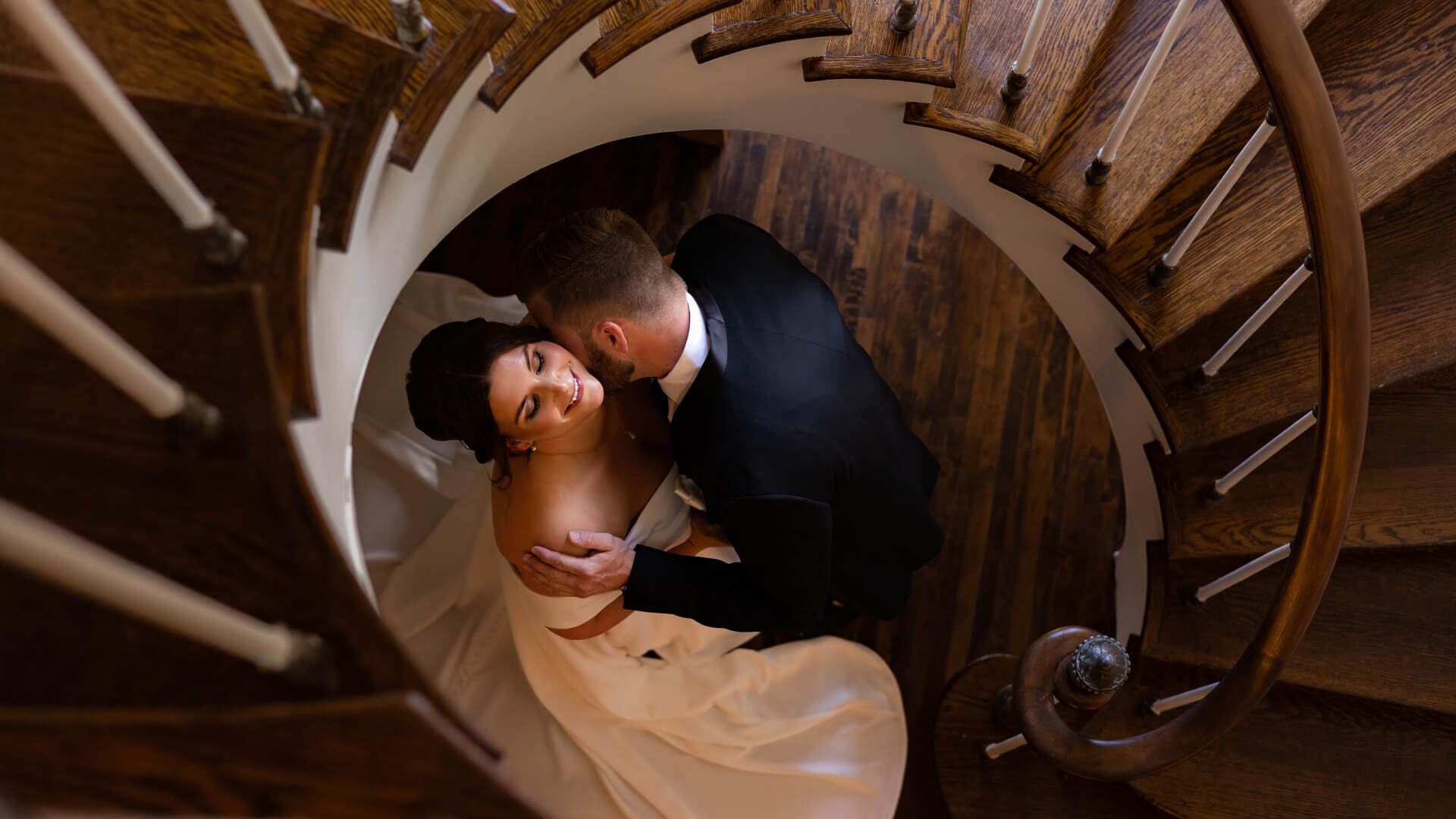 A Michigan bride in a white dress and a groom in a black suit embrace at the bottom of a wooden spiral staircase, viewed from above. The scene is warmly lit and captures an intimate moment.