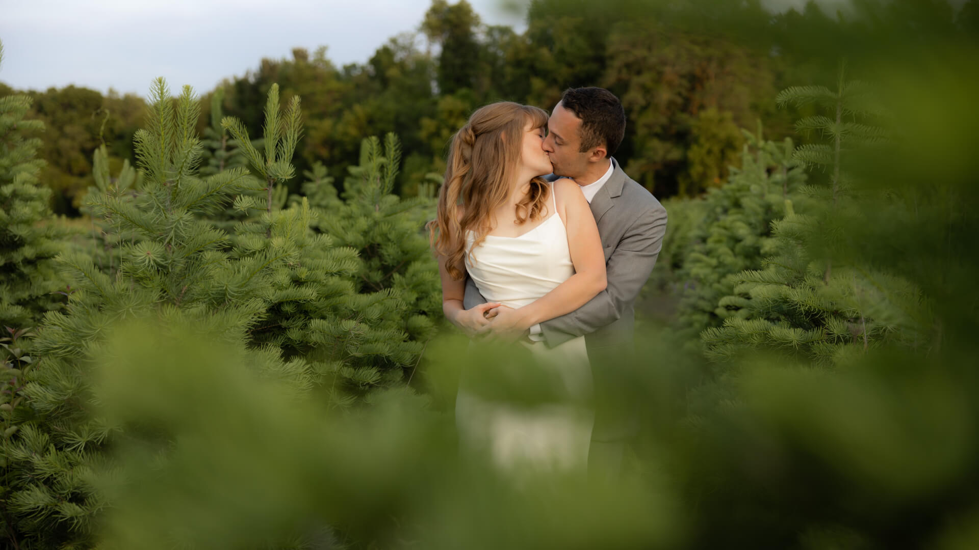 A Michigan couple in wedding attire embraces and kisses while standing among green pine trees in an outdoor setting, with trees and sky in the background.