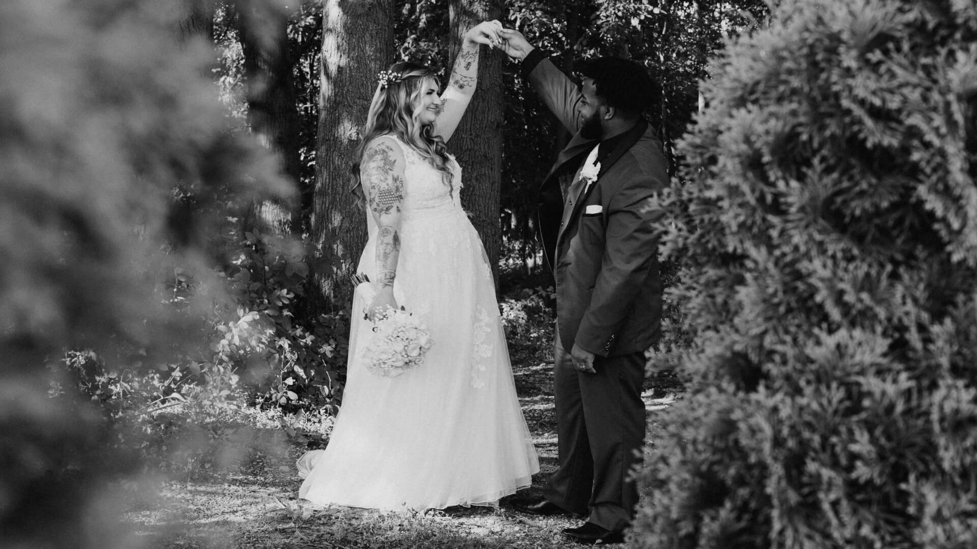 A Michigan bride in a white dress and a groom in a suit dance together outdoors, surrounded by trees and foliage. The bride holds a bouquet and the groom lifts her hand. The scene is in black and white.