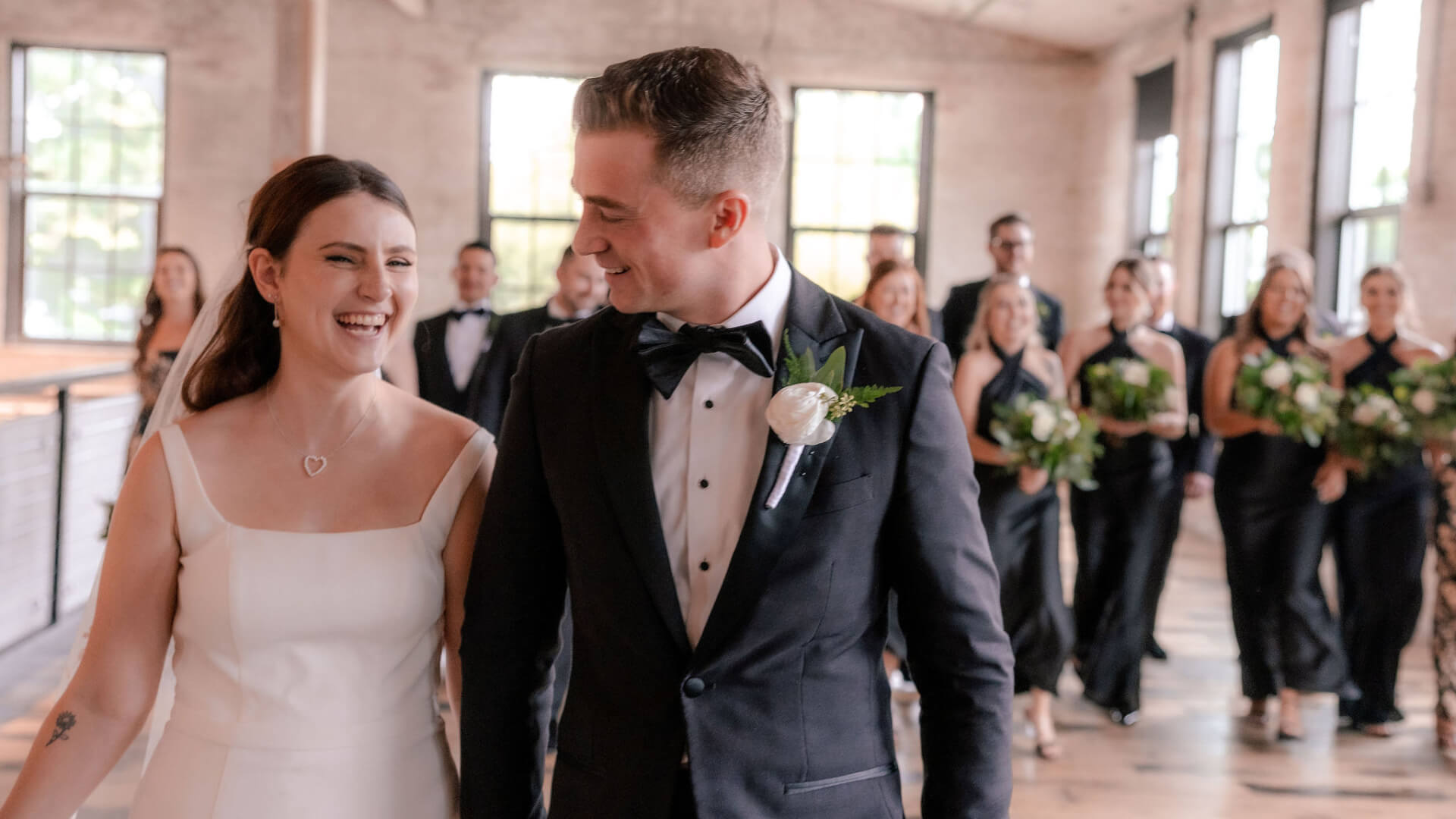 A Michigan bride and groom walk indoors, smiling at each other. The bride wears a white dress and the groom wears a tuxedo. Bridesmaids and groomsmen holding bouquets follow behind them. The scene is well-lit with large windows.