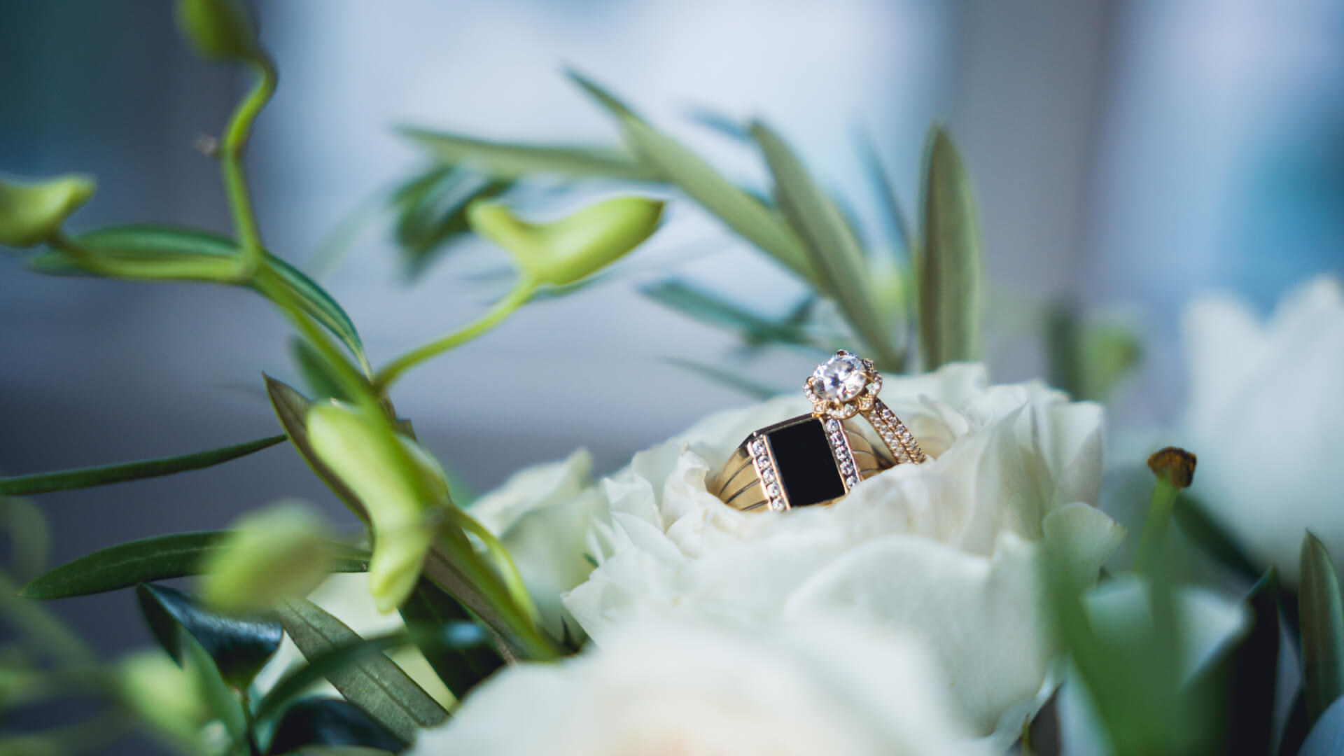 A diamond engagement ring and a gold wedding band rest on white flowers with green leaves in the background.