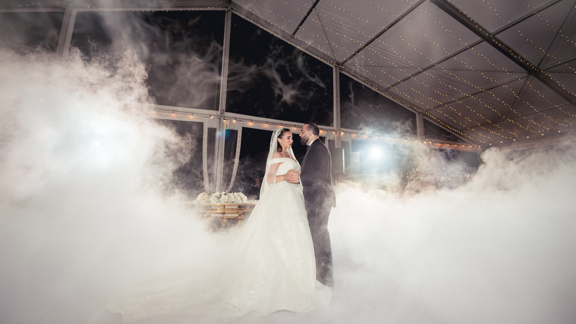 A bride and groom stand together in the center of a misty, fog-filled wedding venue with string lights overhead and a glass ceiling, looking at each other under bright lighting.