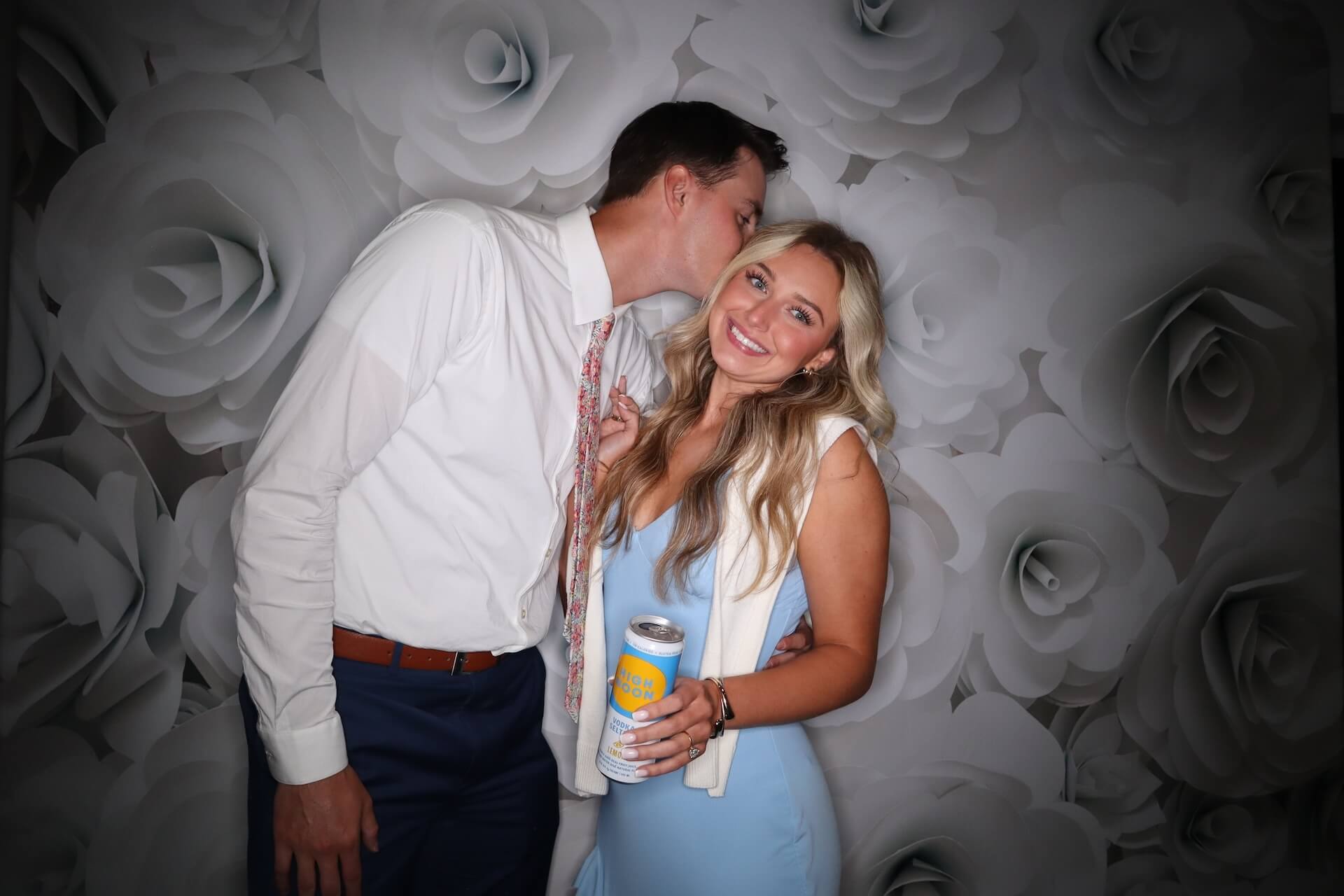 A man in a white shirt and tie kisses a smiling woman in a light blue dress on the side of her head. The woman holds a canned drink. The background features a pattern of large white roses.