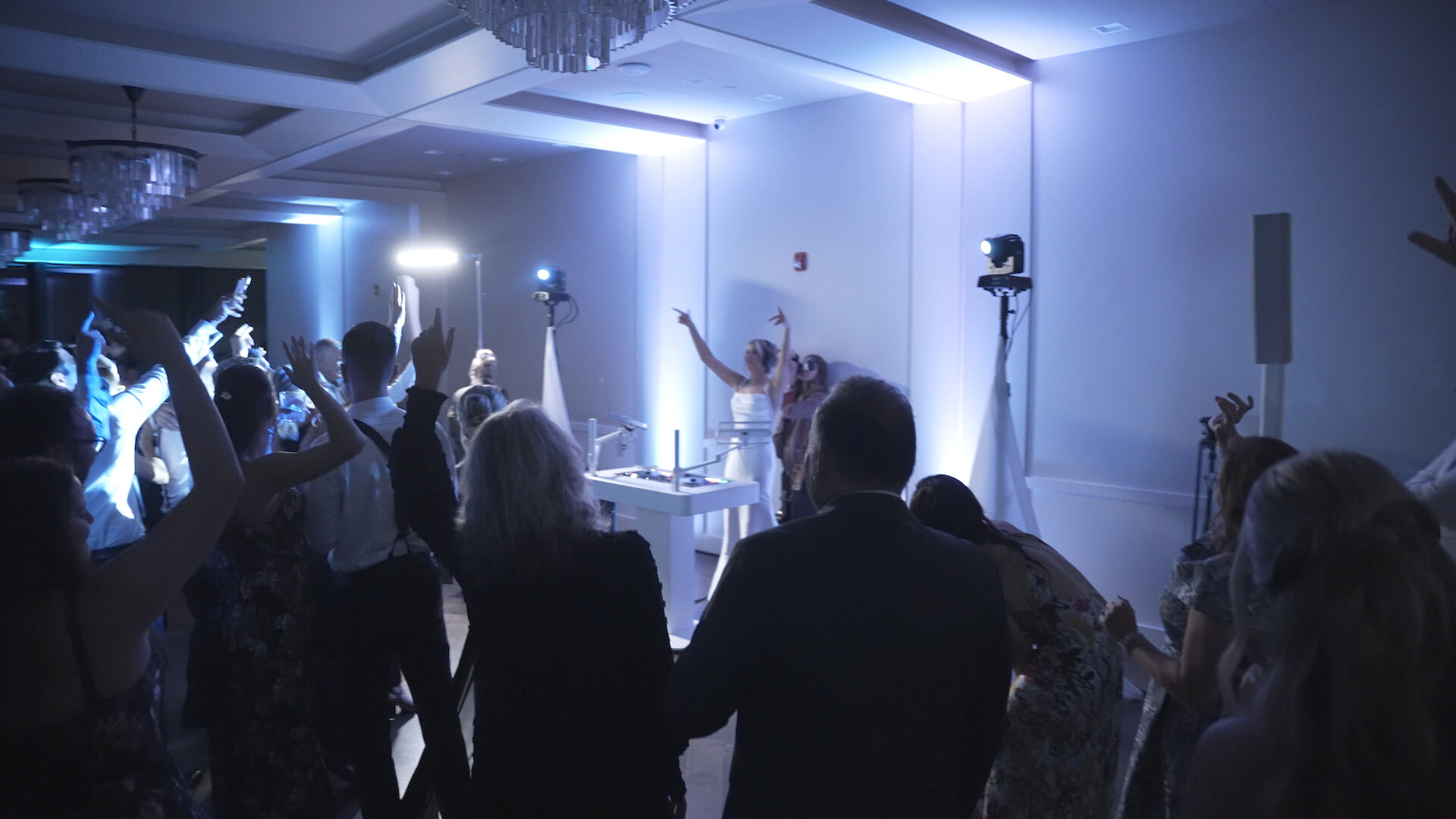 A group of people in formal attire are dancing and raising their hands in a dimly lit room with blue lighting. A DJ stands at a table in the background, and spotlights illuminate parts of the scene.