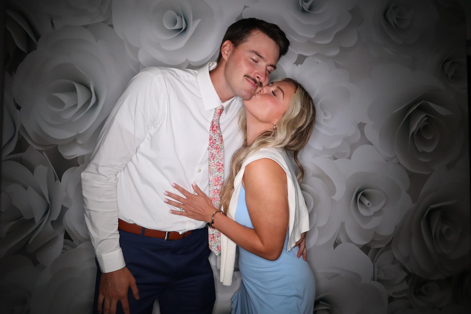 A woman in a light blue dress kisses a man in a white shirt and floral tie on the cheek. They stand in front of a gray and white rose-patterned backdrop, posing closely and smiling with eyes closed.