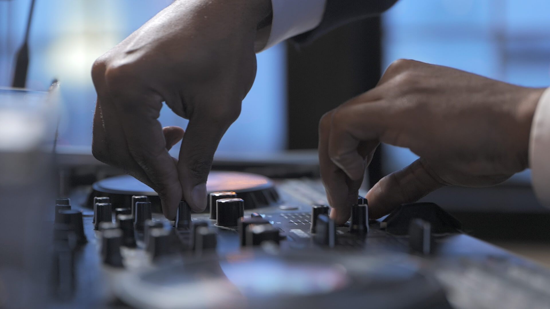 Close-up of a person's hands adjusting knobs and sliders on a professional DJ mixing console, with a blurred background.