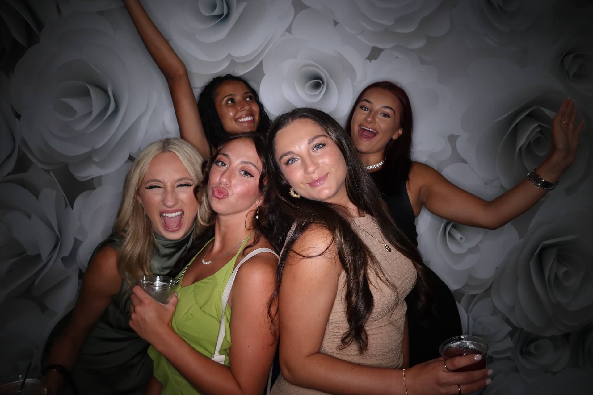 Five women pose together in front of a wall decorated with large, white rose patterns. Some are smiling, others are making playful faces. Several hold drinks and have their arms raised or around each other.