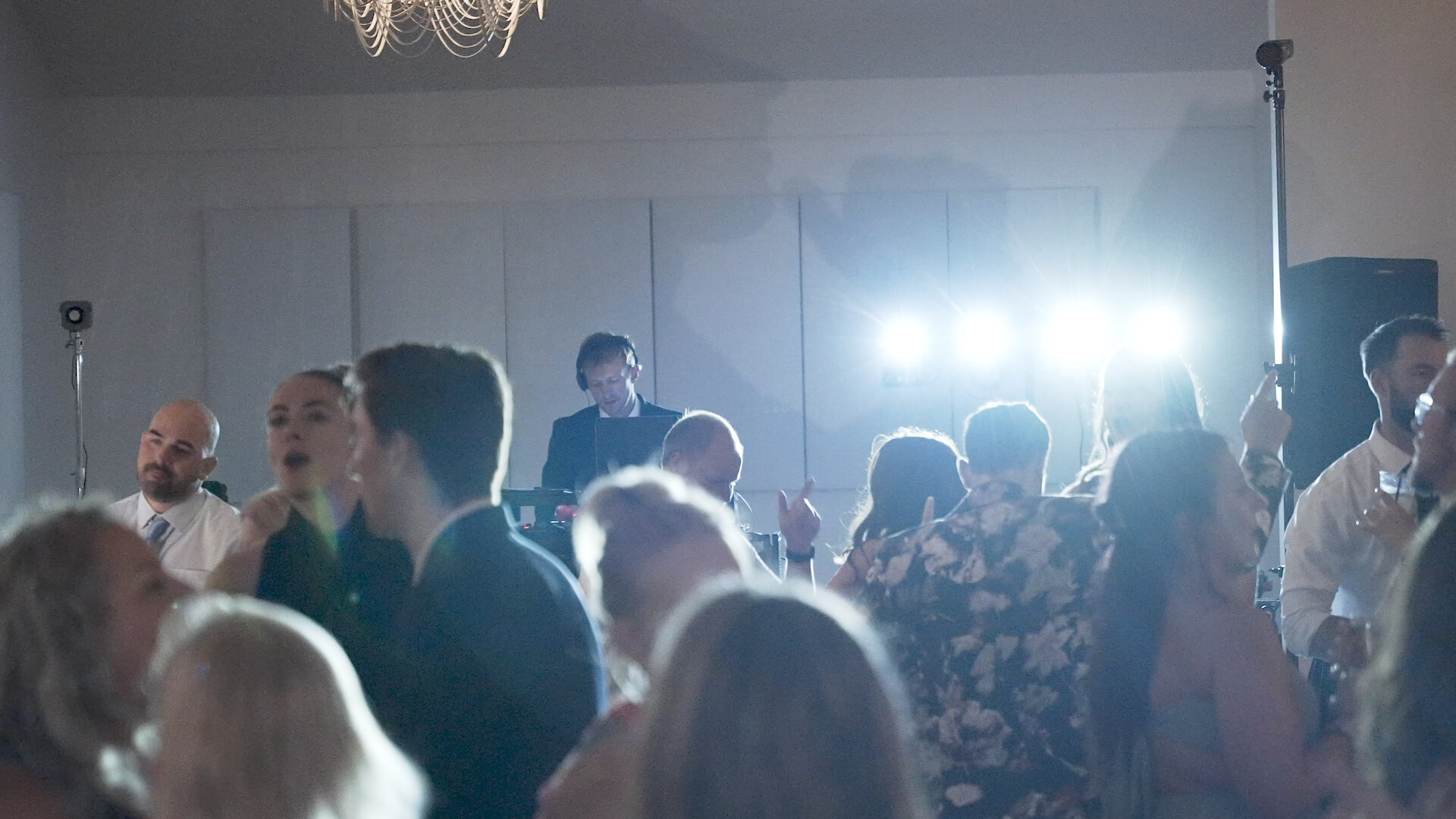 A group of people are dancing at an indoor event with a DJ playing music in the background. Bright lights are shining towards the camera, creating a lively atmosphere. Some individuals are talking while others dance.