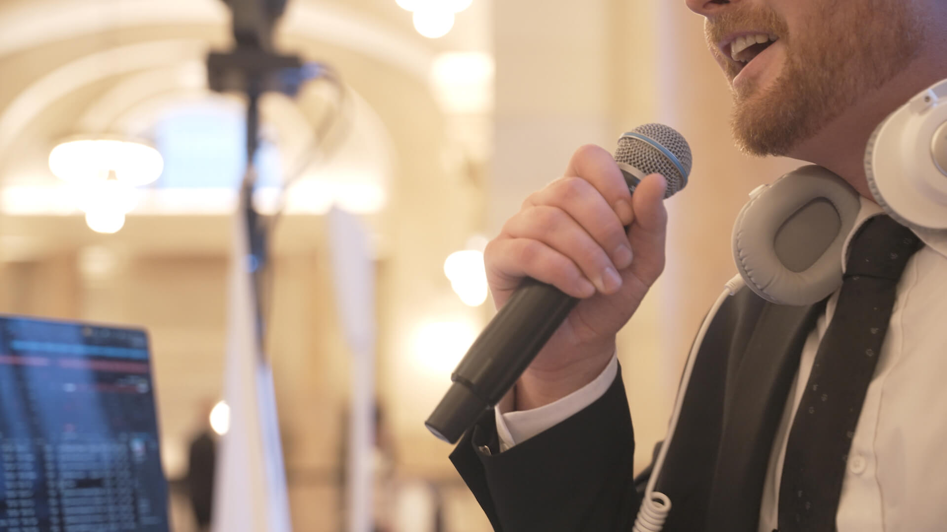 A man in a suit holds a microphone and speaks. He has headphones around his neck and is standing near audio equipment in a well-lit indoor venue. The background is out of focus.