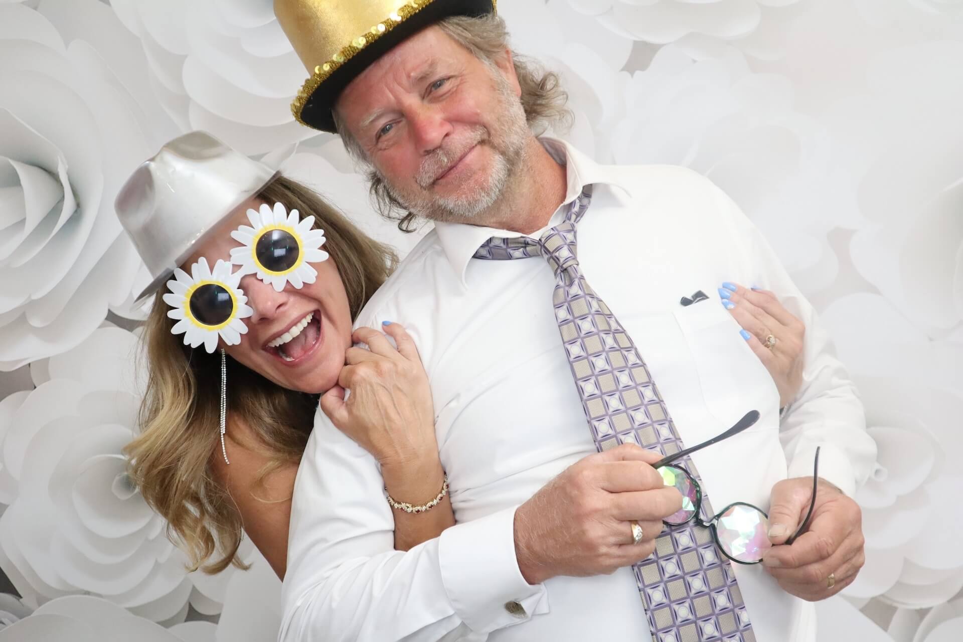 A woman wearing flower-shaped sunglasses and a silver hat stands next to a man in a gold hat and patterned tie. Both are smiling in front of a white floral backdrop. The man is holding a pair of glasses.