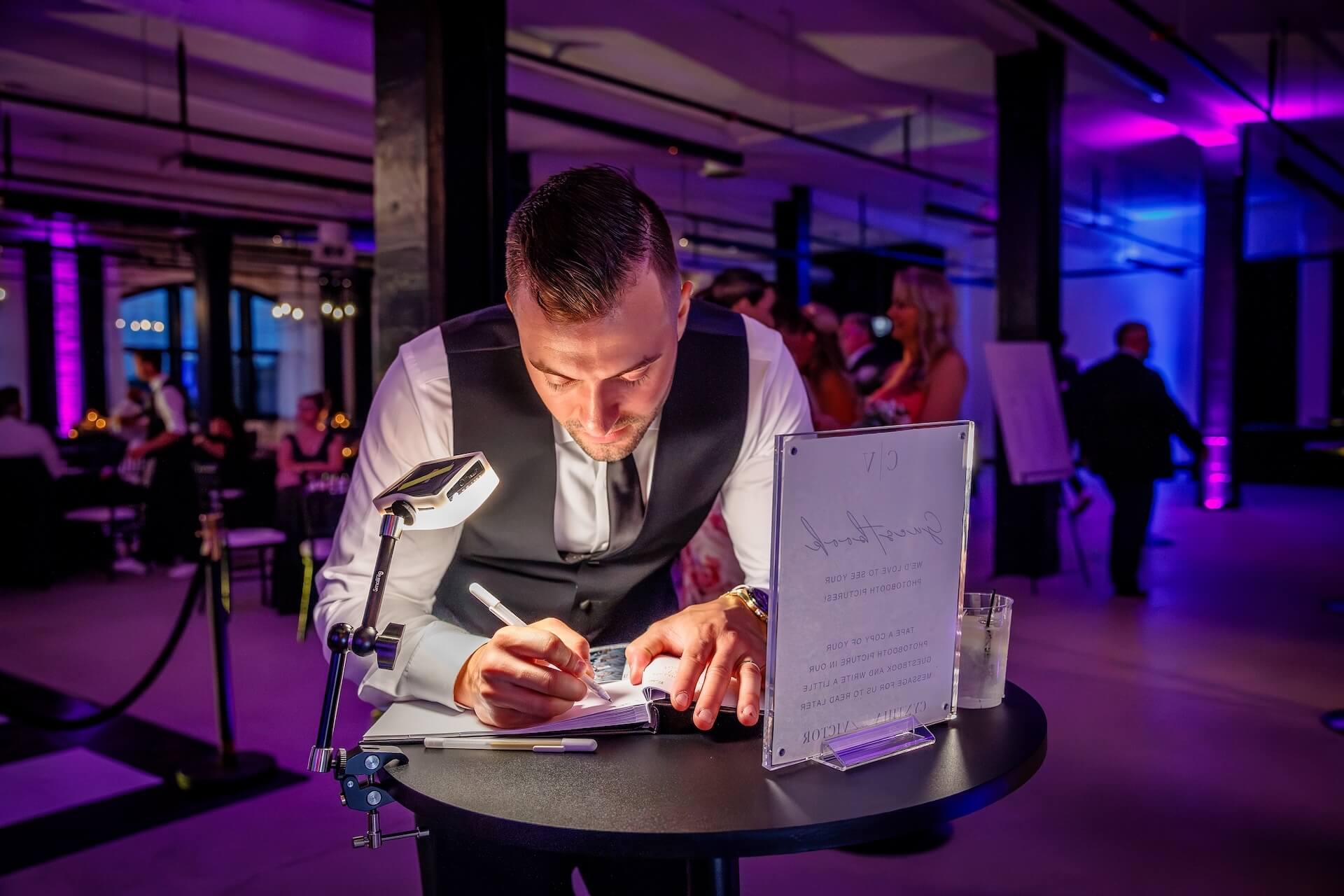 A man in formal attire writes on a notepad at a round table with a drink and a framed sign, in a dimly lit room with purple lighting and people in the background.