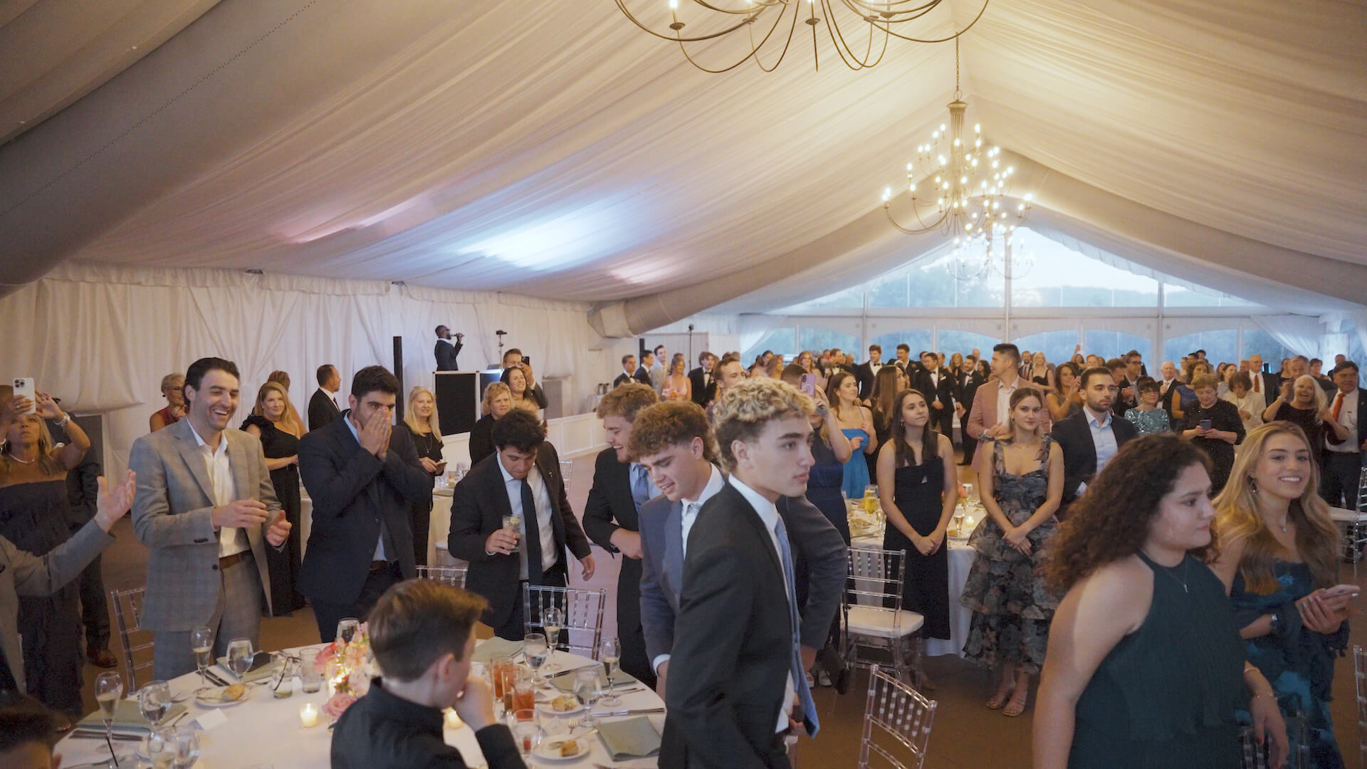A large group of people dressed in formal attire stands and sits at tables in a white tent with chandeliers, attending an event or celebration. Some guests are talking, while others look toward the front.