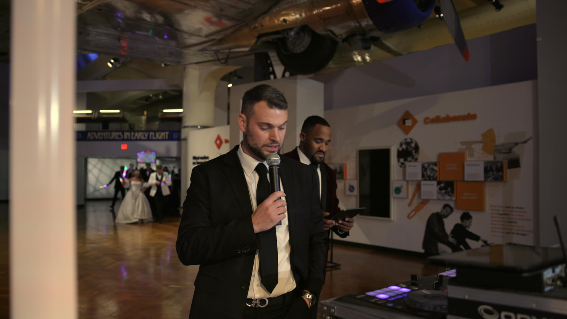 A man in a black suit and tie speaks into a microphone at an indoor event space with another man standing behind him. There are displays on the walls and a wedding couple visible in the background.