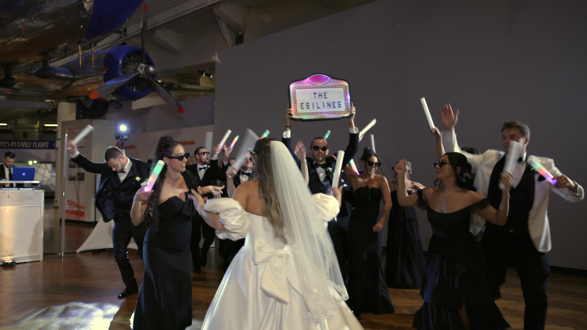 A bride in a white dress and veil dances with guests in black attire, some wearing sunglasses and holding light sticks. One person holds a sign reading "The Estilines." The setting is an indoor venue with a plane visible above.