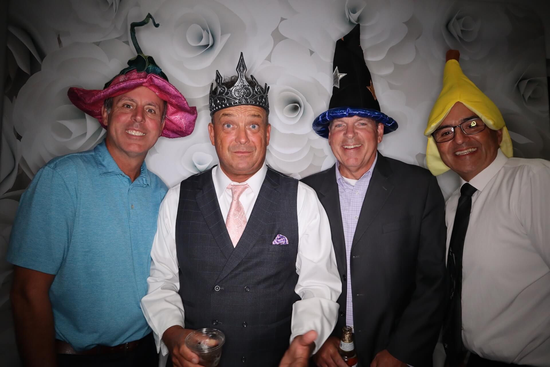 Four men dressed in business attire stand in front of a floral-patterned backdrop, each wearing a whimsical hat: a pink flower, a silver crown, a blue wizard hat, and a yellow banana. Two men hold drinks.