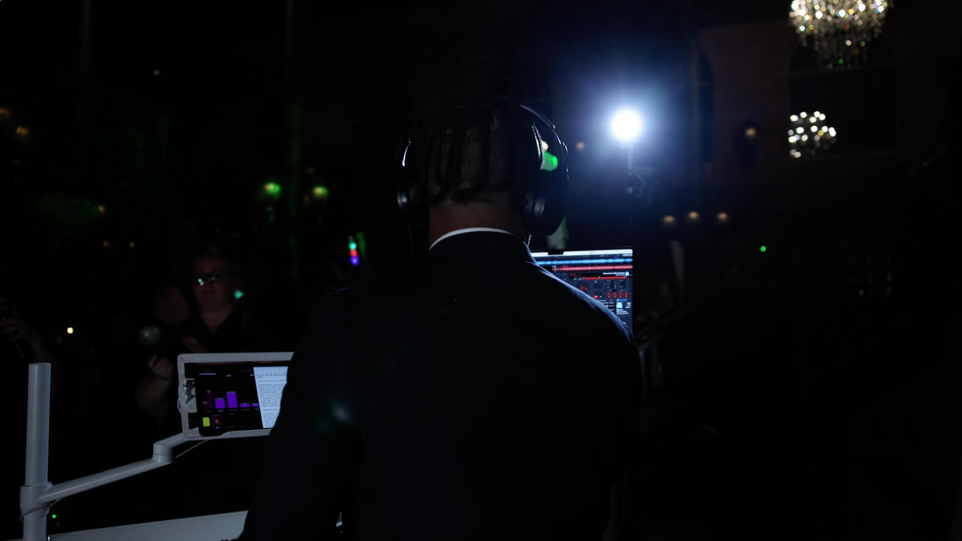 A person wearing headphones stands in front of DJ equipment and computer screens in a dimly lit room, with a bright light source shining from behind.