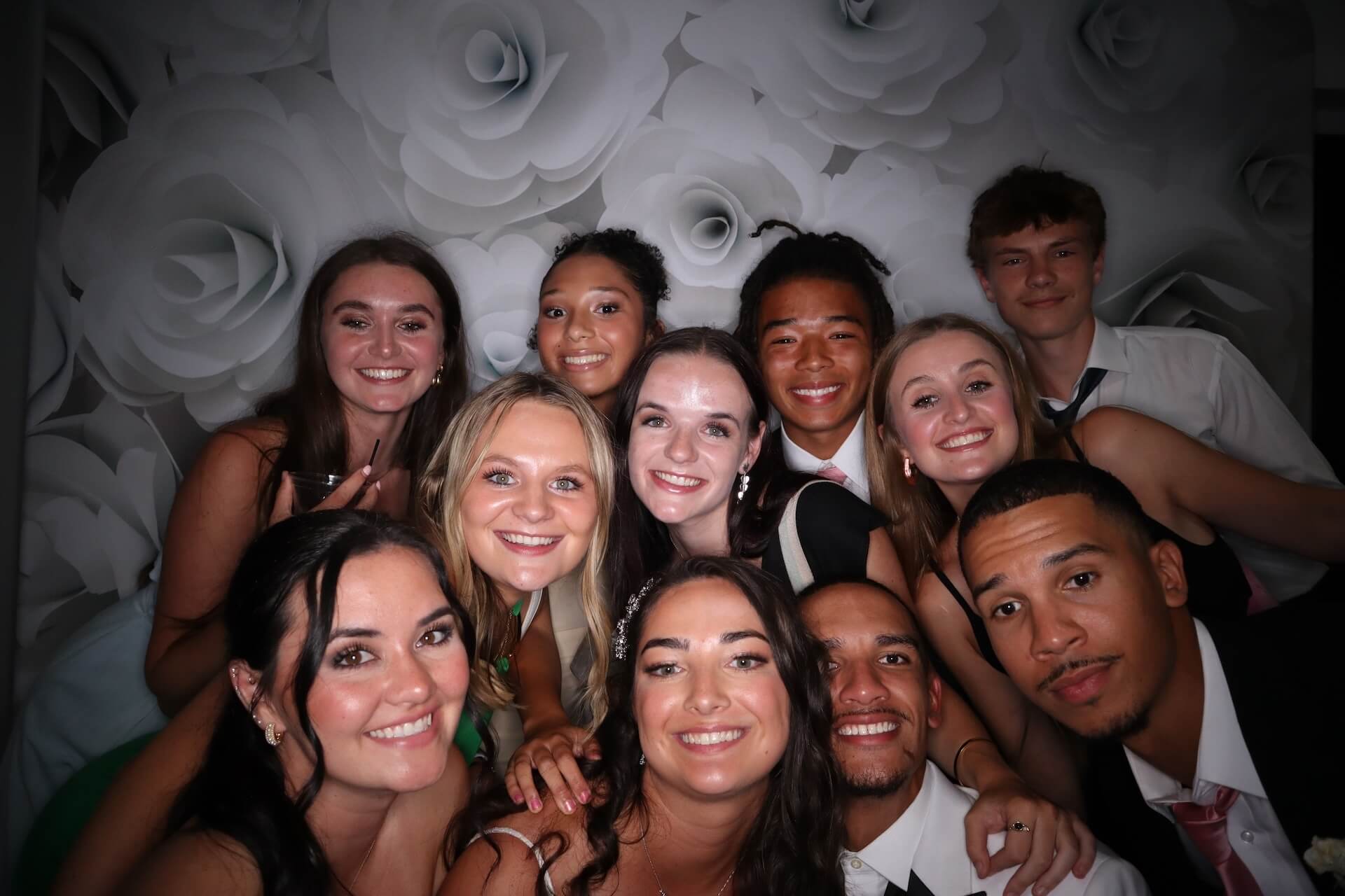 A group of eleven young adults dressed formally smile closely together for a photo in front of a backdrop with large white paper flowers.